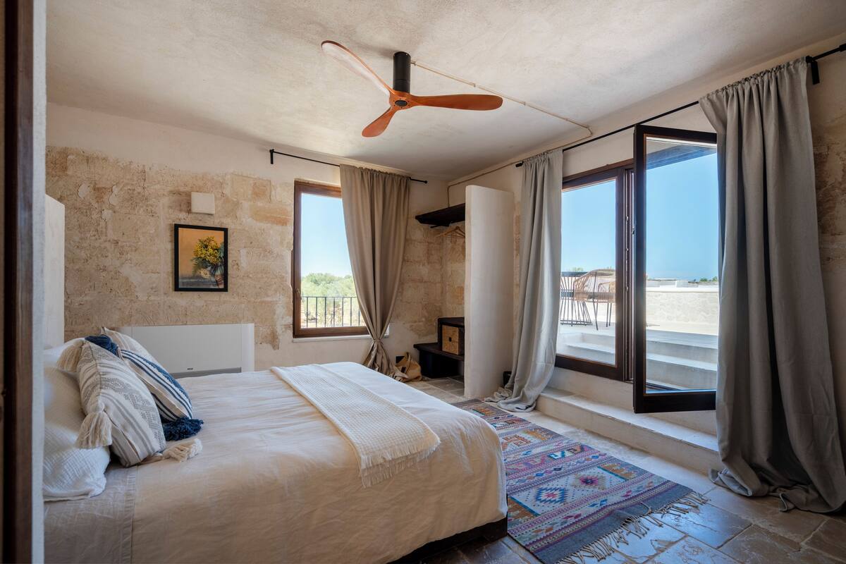Ein Schlafzimmer mit Bett, Deckenventilator, Fenster mit Vorhängen und Blick ins Freie.