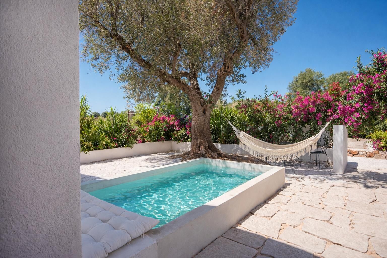 A rectangular pool with a white tiled edge, surrounded by a stone patio. There is a hammock and a chair near the pool, and a tree with pink flowers in the background.