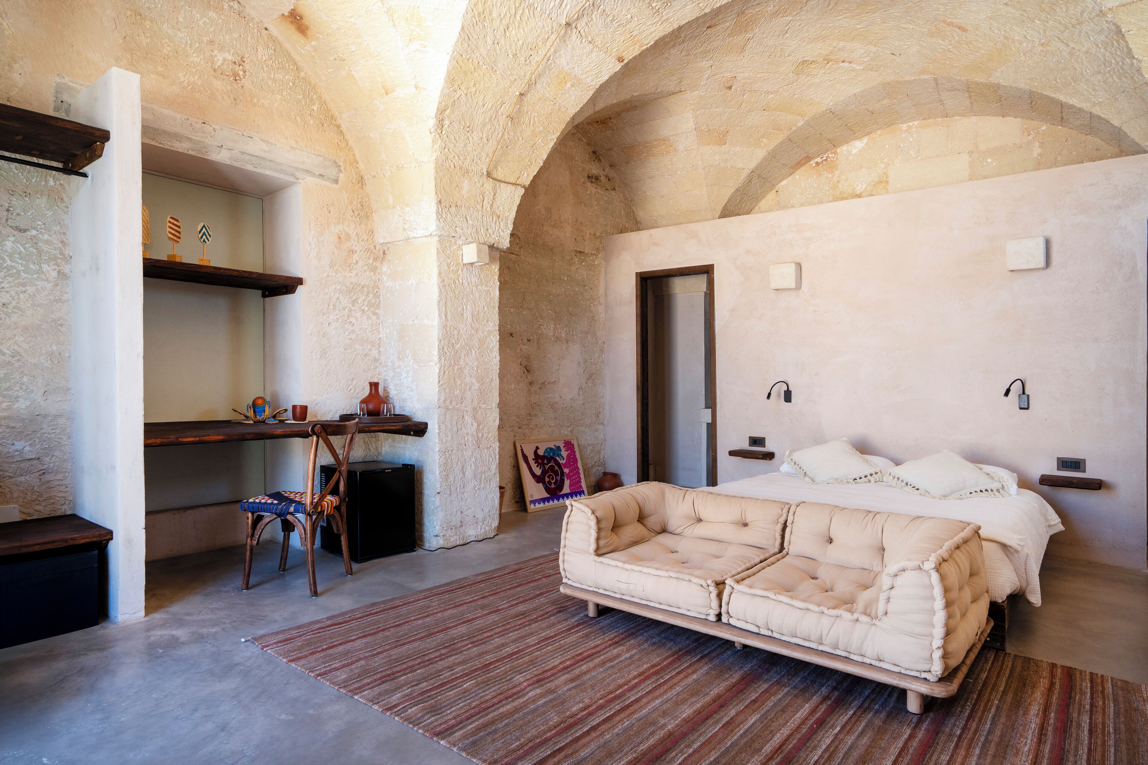 A bedroom with a large bed, a desk, and a chair. There is a door and a shelf with decorative items.