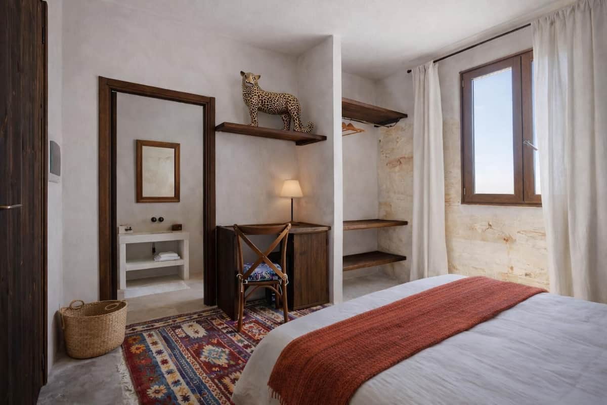 Ein Schlafzimmer mit Bett, Nachttisch, Stuhl, einer Ablage mit einer Leopardenfigur, einer Pendelleuchte und einem Fenster mit Vorhängen.