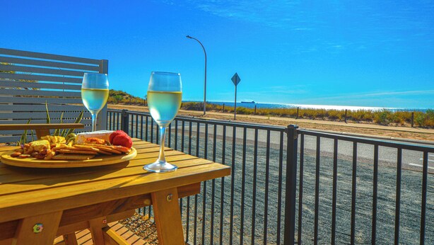 Ein Holztisch mit einem Teller Essen und zwei Gläsern Weißwein, mit Blick auf einen Strand unter blauem Himmel.