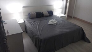 A neatly made bed with a grey bedspread and two pillows, a bedside table with a lamp, and a small white nightstand.