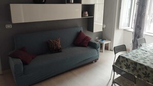 A modern living room with a blue sofa, two pillows, a small side table, a lamp, and a dining table with chairs.