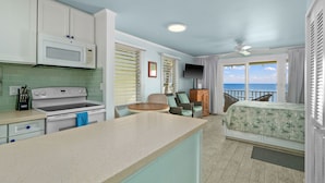 A compact kitchen with a microwave, stove, and dining area. There is a TV mounted on the wall and a balcony with a view of the ocean. - Oceanfront Studio with Panoramic Views - Kapa'a Sands 09 (Kapaa)