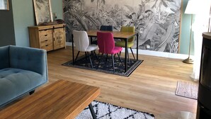 Ein Wohnzimmer mit Couch in Blau, einem Holztisch, einem Esstisch mit bunten Stühlen und einer Wandmalerei von tropischer Vegetation.