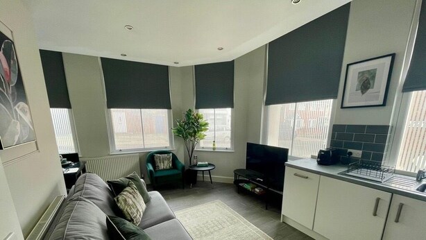 Una sala de estar moderna con un sofá gris, una mesita auxiliar, un televisor y una zona de cocina con armarios y un lavamanos.