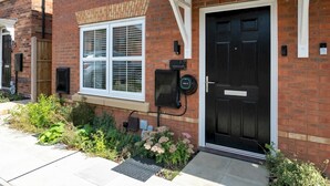 Una casa de ladrillo con puerta principal negra, una ventana con persianas y un pequeño jardín con flores rosas.