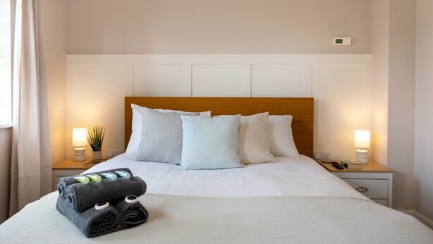 Una cama bien hecha con sábanas blancas y un cabecero de madera, dos lámparas de noche, una mesita de noche con una planta y una toalla doblada sobre la cama.