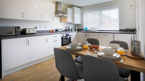 Una cocina moderna con armarios blancos, una mesa de madera dispuesta para el desayuno y una ventana con persianas.