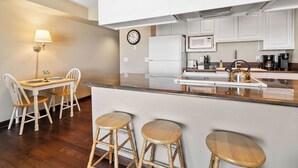A modern kitchen with white cabinets, a stainless steel refrigerator, a microwave, and a coffee maker. There is a wooden dining table with two chairs and three stools. A wall clock is visible on the wall. - Beautiful Luxury Bay View Condo! (Lincoln City)