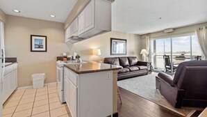 A modern kitchen with white cabinets, a central island, and a dining area with a dark leather sofa and a dining table. - Beautiful Luxury Bay View Condo! (Lincoln City)