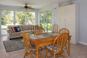 Un espace repas avec une table et des chaises en bois, un ventilateur de plafond, un canapé avec des coussins et une vue sur la verdure grâce à de grandes fenêtres.
