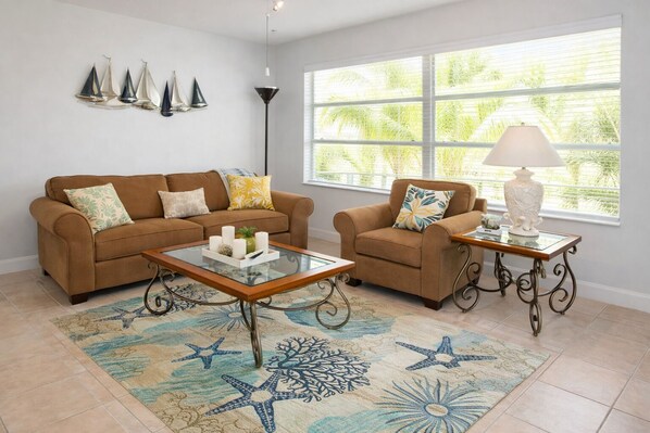 Un salon avec un canapé marron, un fauteuil assorti, une table basse en verre, un tapis à motifs, un lampadaire et une fenêtre avec des stores.