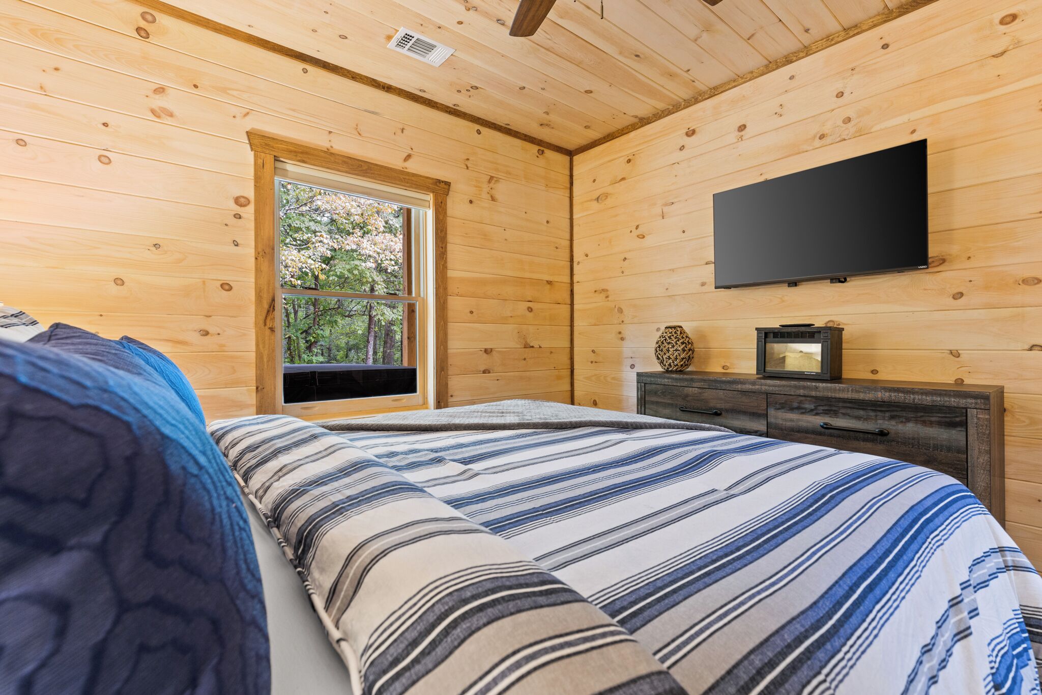 A wooden-paneled bedroom with a bed, a dresser, a flat-screen TV, and a window with a view of trees.