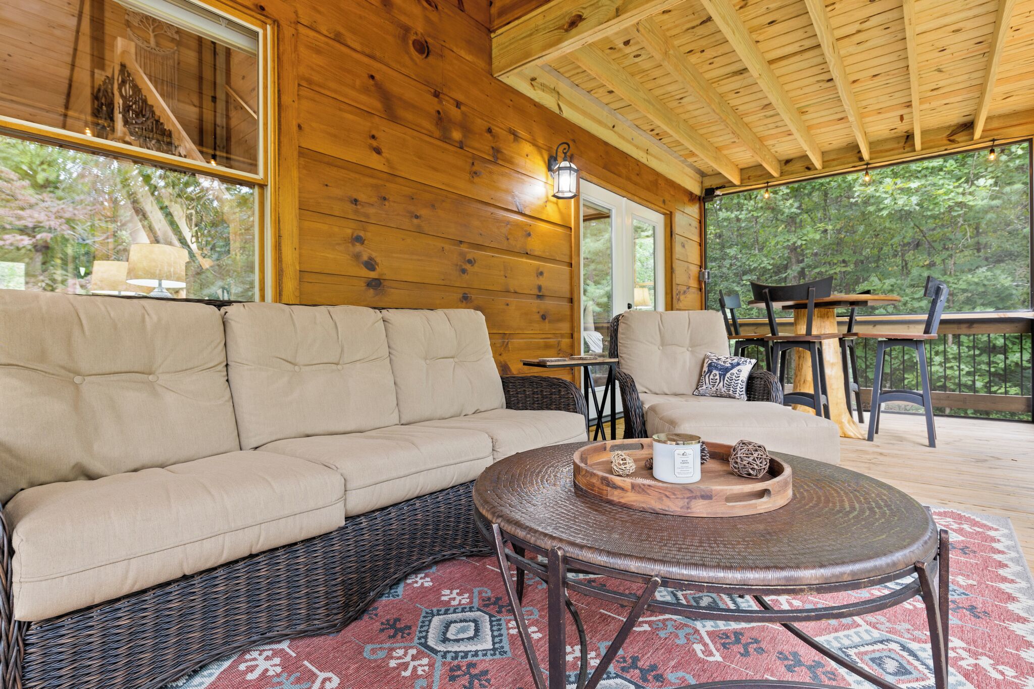 Ein Interieur einer Holzhütte mit einem beigefarbenen Ecksofa, einem runden Korbstuhl-Tisch und einem Esstisch mit Stühlen.