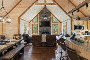 A log cabin interior with a stone fireplace, wooden furniture, and a kitchen area. - Deerly Blissed, Blue Ridge (Blue Ridge)