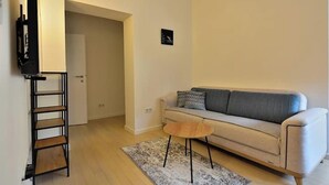 A living room with a sofa, a coffee table, a TV on a stand, and a door leading to another room. - BASC Modern 2 (Sarajevo)