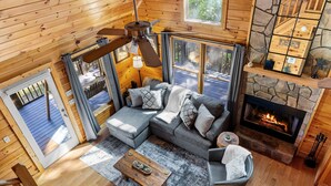 A cozy cabin interior with a stone fireplace, a wooden ceiling fan, and a large window overlooking a deck.