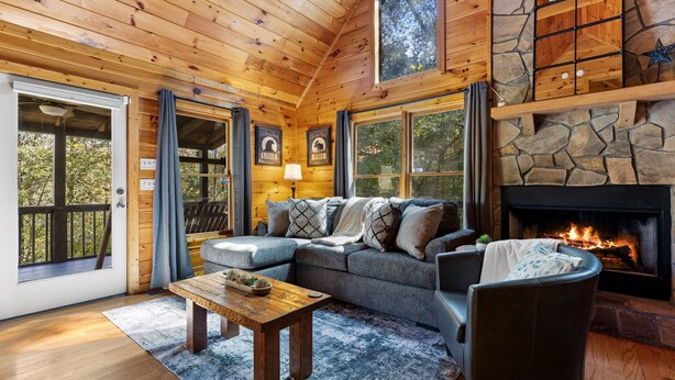 A cozy cabin interior with a stone fireplace, wooden ceiling, and a large window overlooking the outdoors.