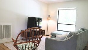A living room with a grey sofa, a wooden chair, a flat-screen TV, a floor lamp, and a large window. - Quiet home tucked between Crystal River & Rainbow River (Crystal River)
