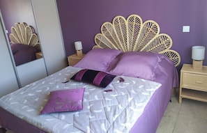 Ein Schlafzimmer mit einem Bett, violetter Bettwäsche, einem Blattmuster-Kopfende, zwei Nachttischen mit Lampen und einem Spiegel.
