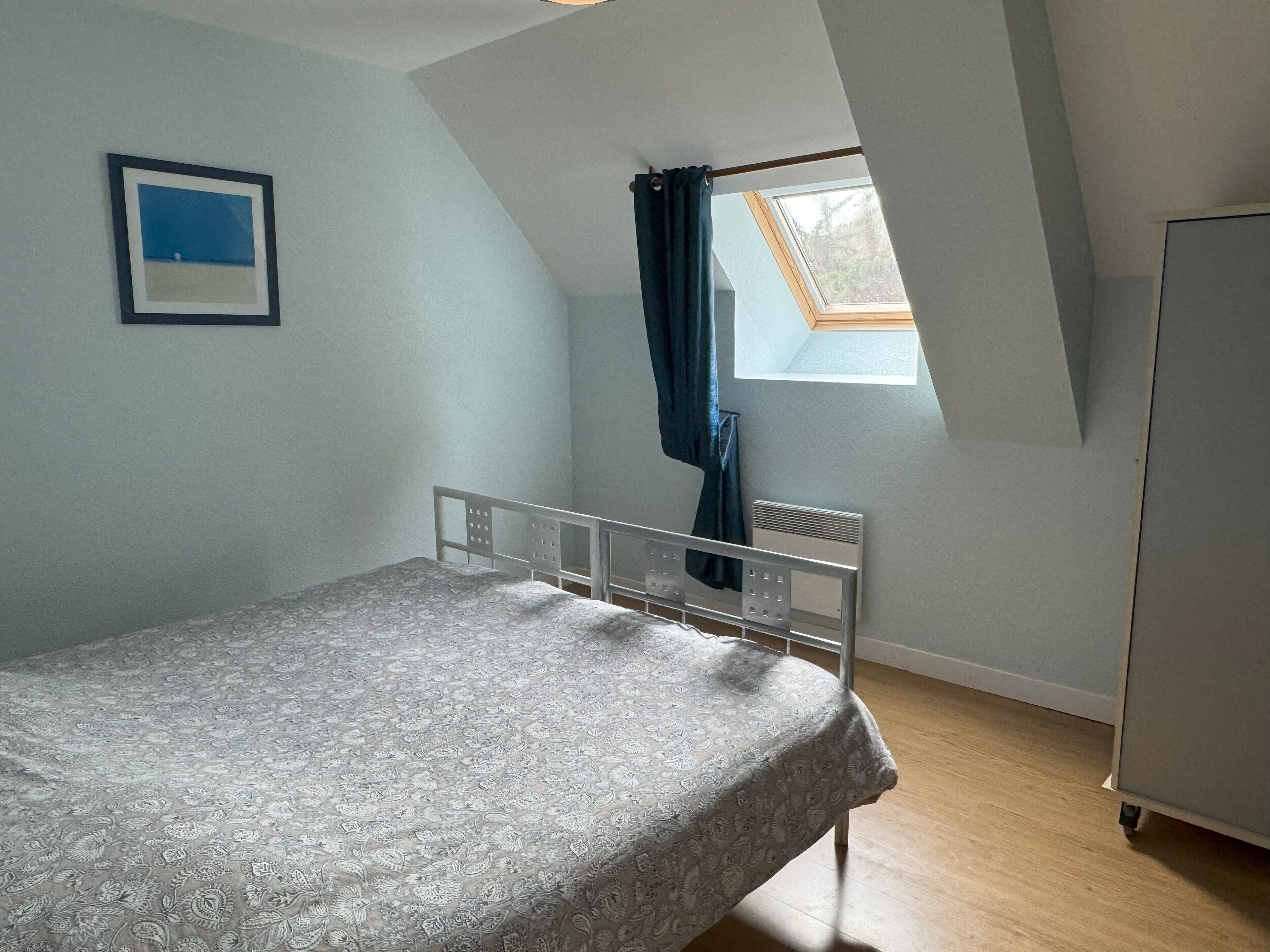 Une chambre avec un lit simple, une fenêtre avec un rideau, un radiateur et un tableau encadré au mur.