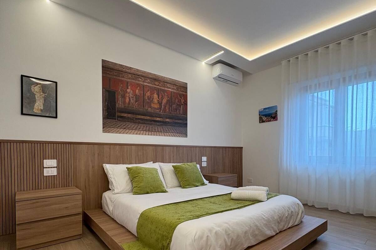 A modern bedroom with a wooden bed frame, a nightstand, a wall-mounted air conditioner, and a large window with sheer curtains.