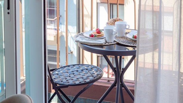 Un balcon avec une table ronde préparée pour le petit-déjeuner, avec des crêpes, des fraises et un croissant. On y trouve deux tasses blanches et un set de table en fibres tressées.