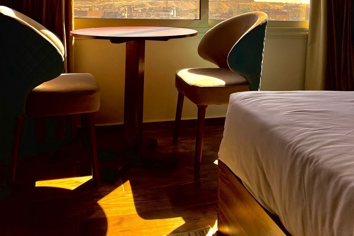 Habitació d'hotel amb un llit, dues cadires, una taula petita i un gran finestral que ofereix una vista de les piràmides.