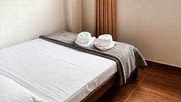 Un letto singolo con lenzuola bianche, una coperta grigia e due cappelli bianchi appoggiati di lato.