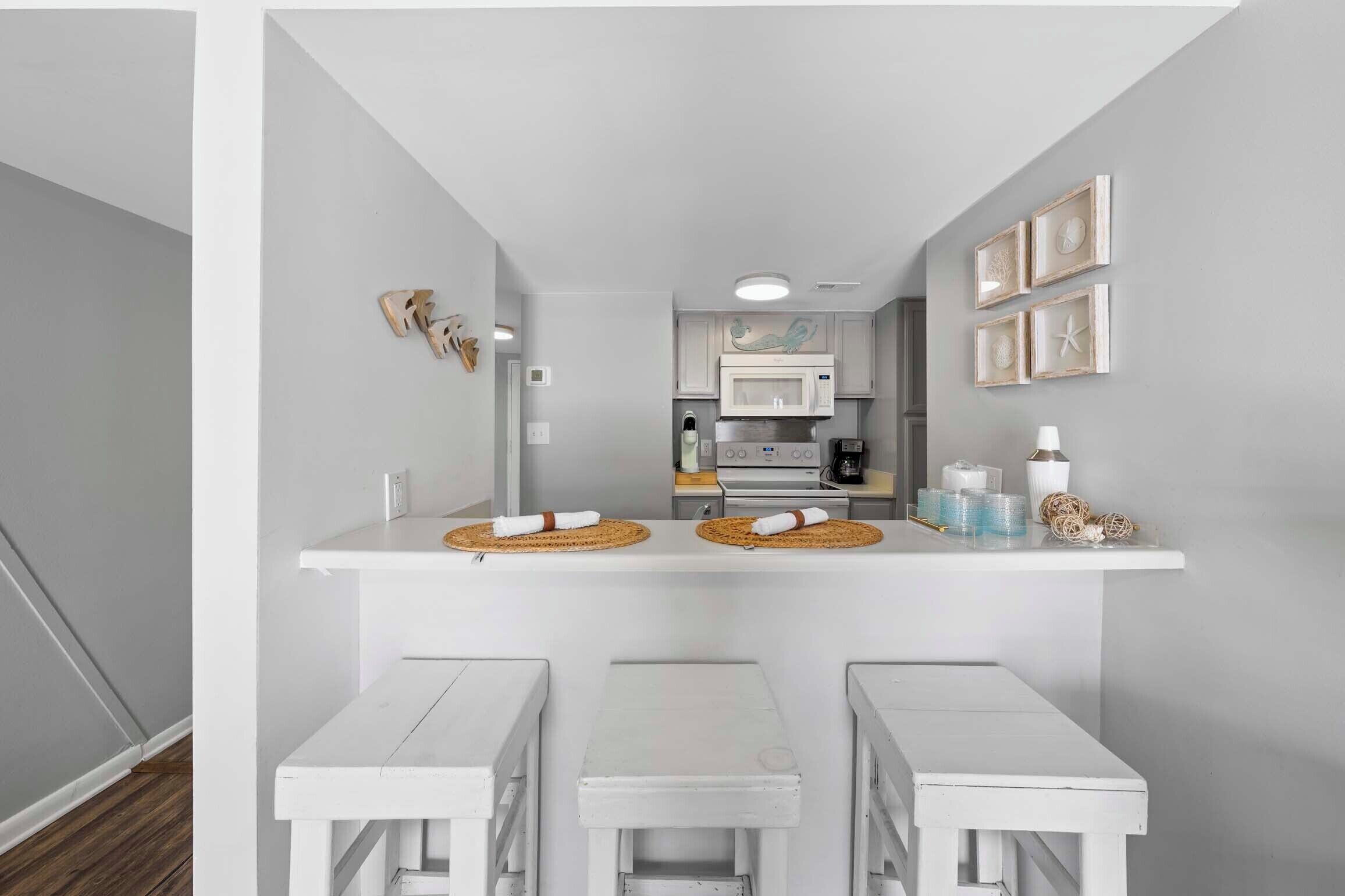 A compact kitchen with white cabinetry, a microwave, and a stove. There are two white tables with woven placemats and a small shelf with decorative items.