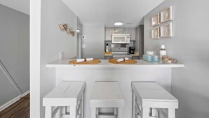 Una cocina compacta con armarios blancos, un microondas y una estufa. Hay dos mesas blancas con individuales tejidos y un pequeño estante con adornos.