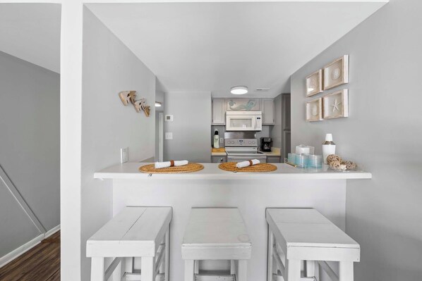 A compact kitchen with white cabinetry, a microwave, and a stove. There are two white tables with woven placemats and a small shelf with decorative items. - 🌊🌴Sugar Beach 364 | Cozy Gulf-View Condo | Steps to Beach + Pools & Tennis (Orange Beach)