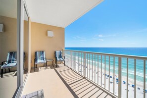Un balcon avec deux chaises et une petite table, donnant sur une plage où l’on peut voir des gens et des parasols.