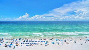 A beach with numerous blue umbrellas and sun loungers, a clear blue sky, and the ocean in the background.
