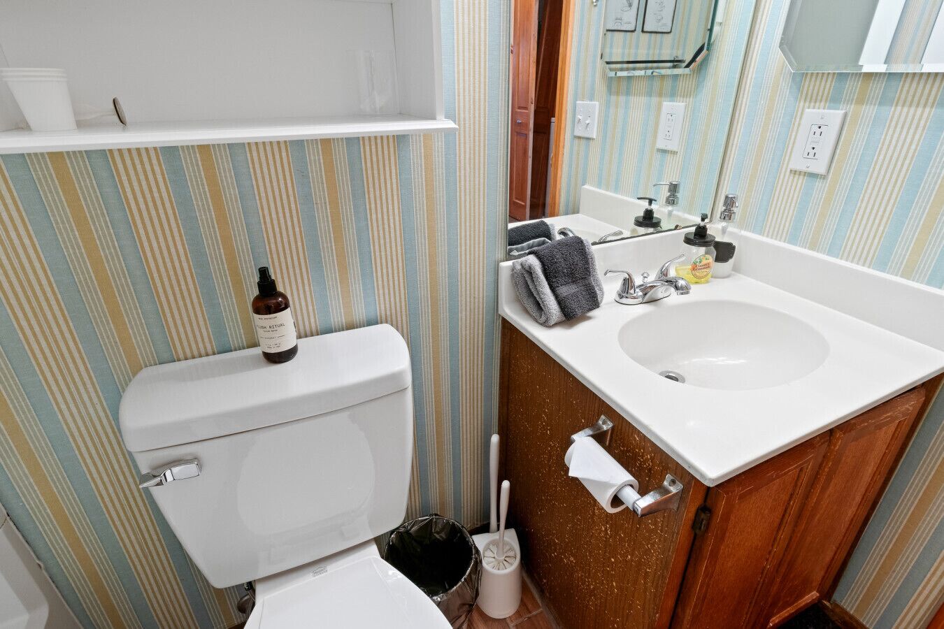 Ein Badezimmer mit gestreifter Wand, einer weißen Toilette, einem Waschbecken mit Spiegel darüber und einem Regal über der Toilette.