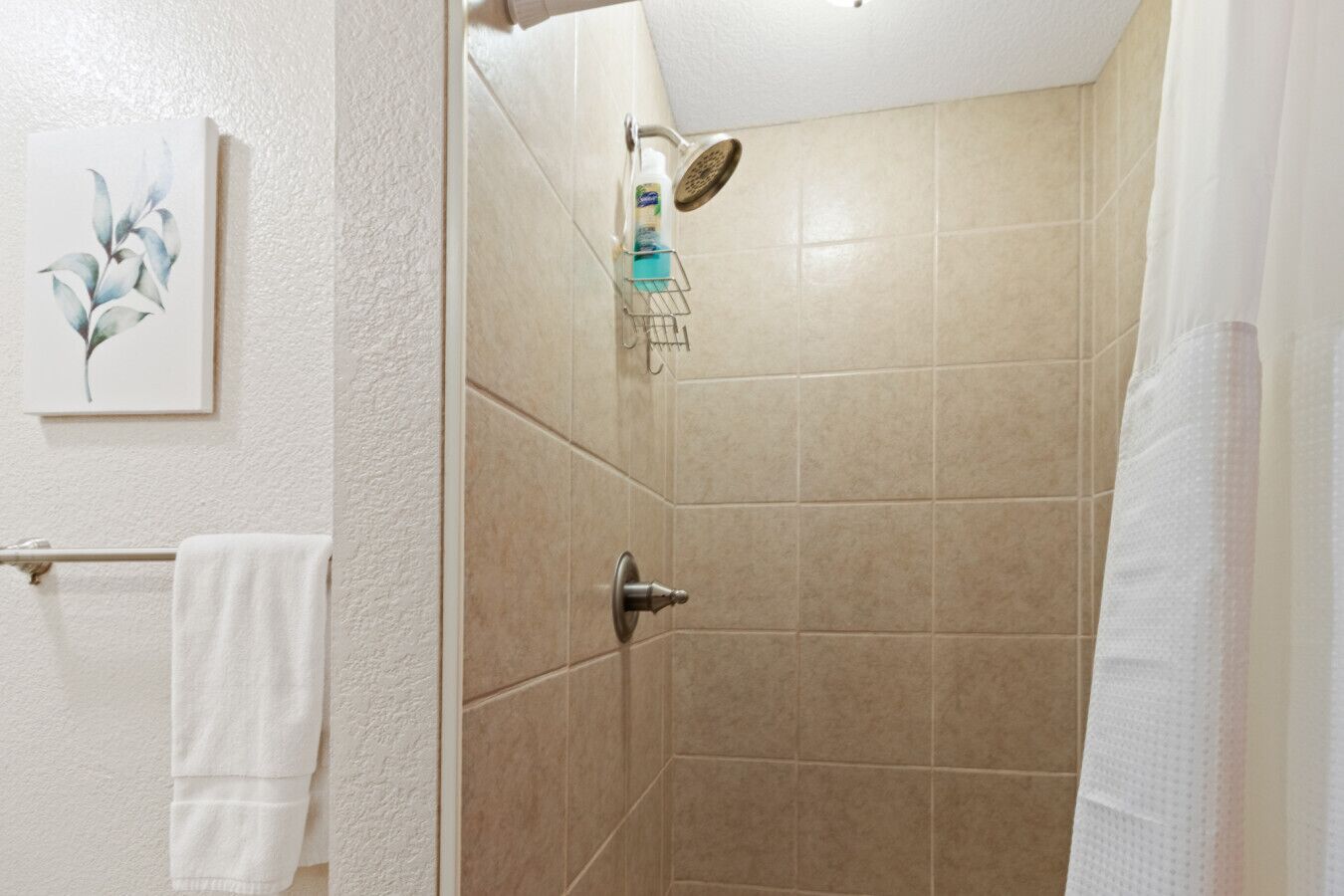 A bathroom with beige tiles, a shower with a handheld showerhead, a towel rack, and a framed picture on the wall.