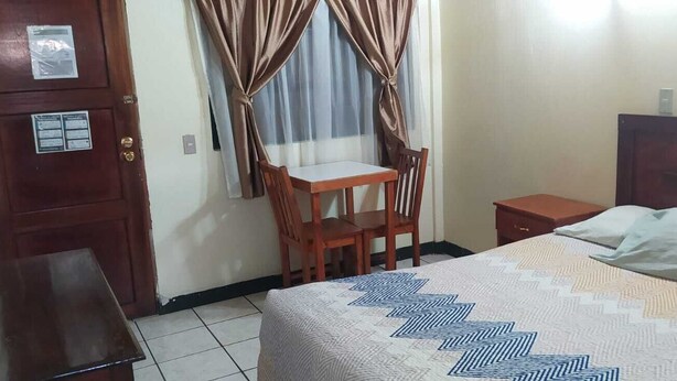 A bedroom with a bed, a wooden nightstand, a chair, a desk, and a window with curtains.