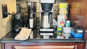 Una cocina con cafetera, licuadora y varios electrodomésticos. Hay una encimera con tabla de cortar y una toalla. Los armarios son de madera con dos pomos.