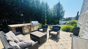 Un patio al aire libre con suelo de losas de piedra, mobiliario de mimbre, una parrilla empotrada y luces colgantes.