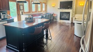 Una cocina moderna con una isla central, taburetes de bar, un comedor con mesa y sillas, una chimenea y un televisor grande montado en la pared.