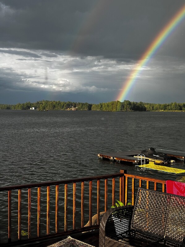 A lakeside view with a wooden dock, a lounge chair, and a distant house.