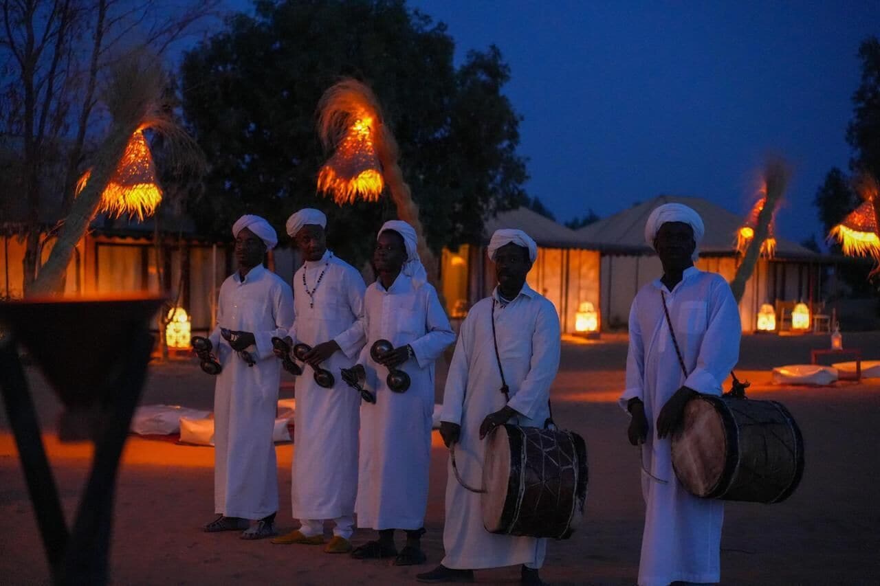 Un grupo de personas con vestimenta tradicional posa en un entorno exterior con una construcción de techo de paja e iluminación cálida.