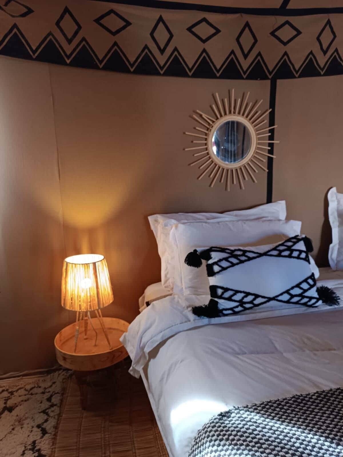 A bedroom with a bed, a round mirror with a sunburst frame, a bedside lamp, and patterned pillows.