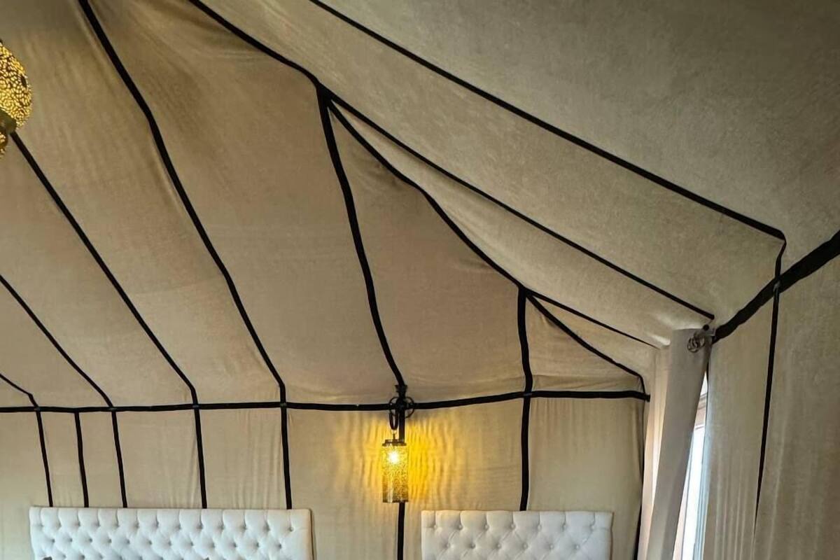 Un dormitorio tipo tienda de campaña con dos camas, cada una con cabecero capitonado y ropa de cama blanca. Se ve una mesita de noche con lámpara en un lateral.