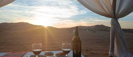 A desert view with a table set for two, featuring wine glasses, a bottle, and bowls of snacks.