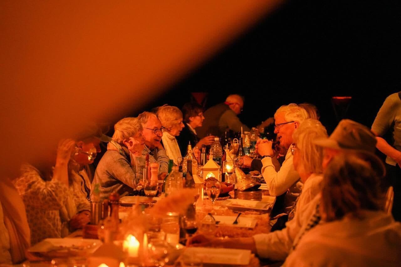 Un groupe de personnes est assis autour d’une table, devant divers bouteilles et verres, ce qui laisse supposer qu’il s’agit d’une réunion sociale ou d’un repas.