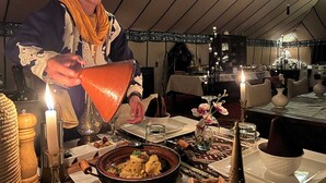 En person som häller upp en drink i ett glas vid ett bord med ett ljus, omgiven av olika rätter och en tältliknande inredning.