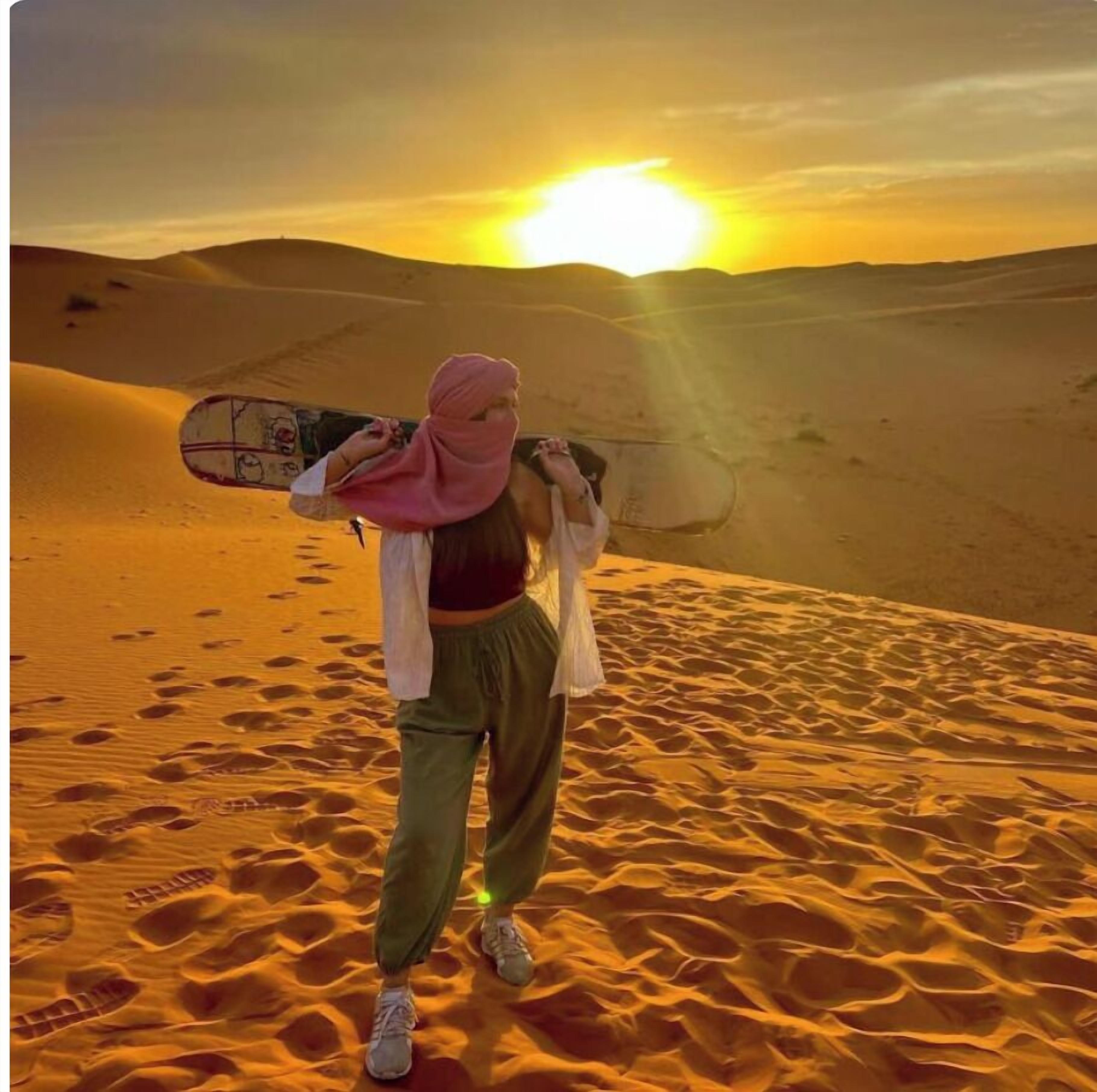 A person standing on a sandy dune with a sunset in the background.