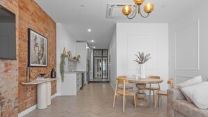 A modern living room with a brick accent wall, a dining area with a round table and chairs, and a sofa with cushions. - Avenue Apartment 1 - luxe Suite -
Inner City - Free Parking (Launceston)